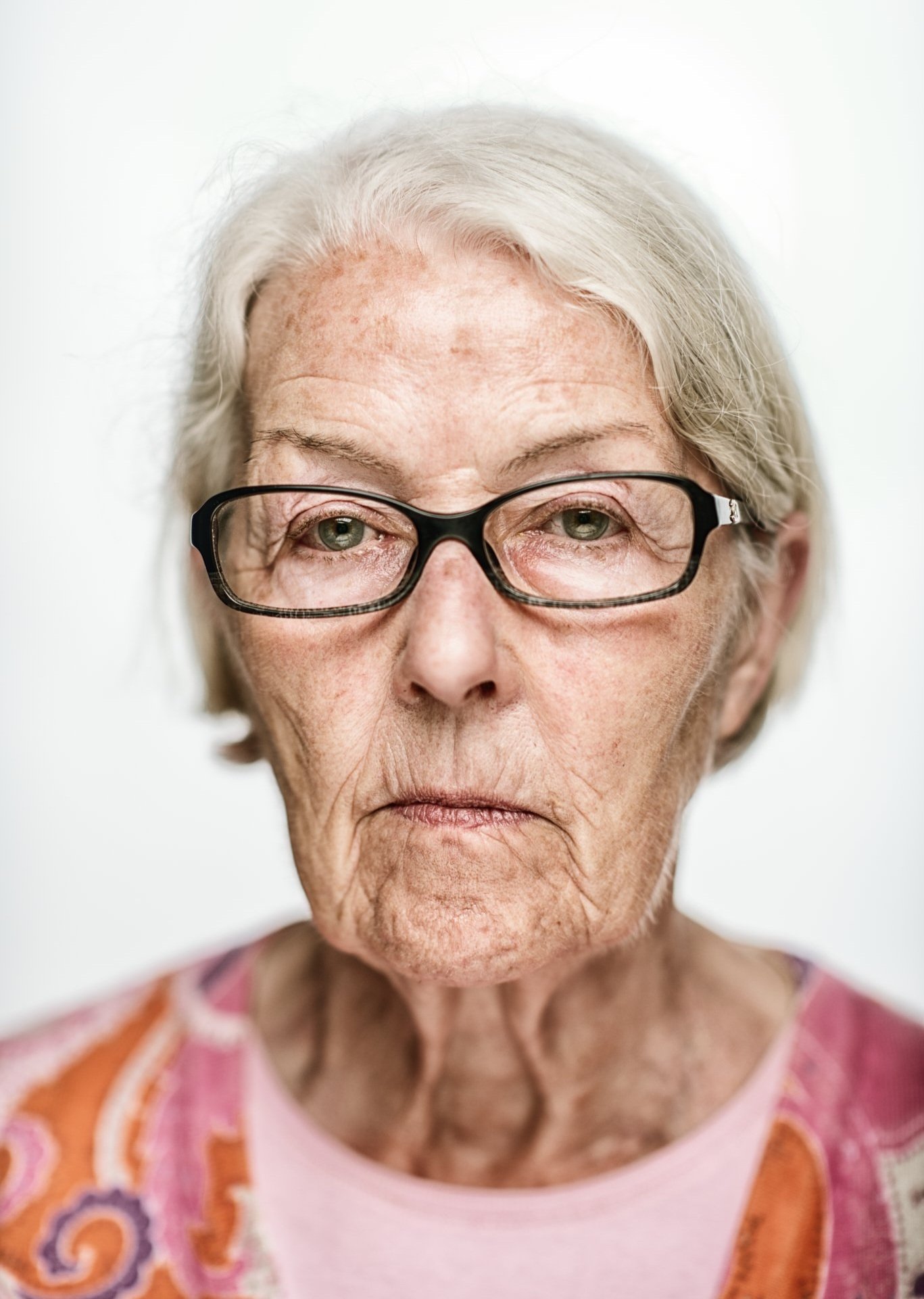 Nahaufnahme eines älteren Menschen mit Brille und grauen Haaren vor einem neutralen Hintergrund. Die Person trägt ein buntes Oberteil.