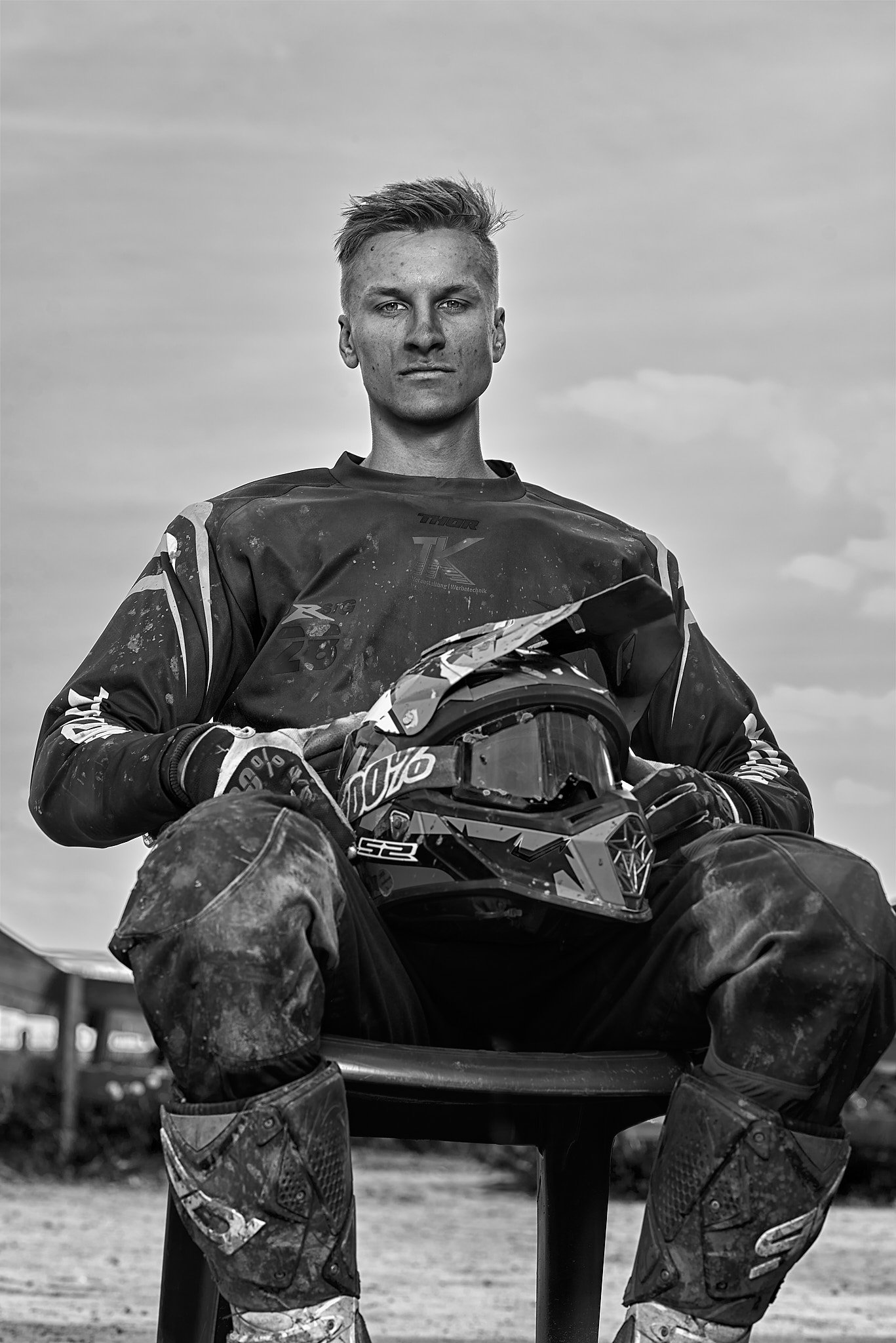 Motocrossfahrer sitzt auf einem Stuhl mit Helm in den Händen, schwarz-weißes Bild.