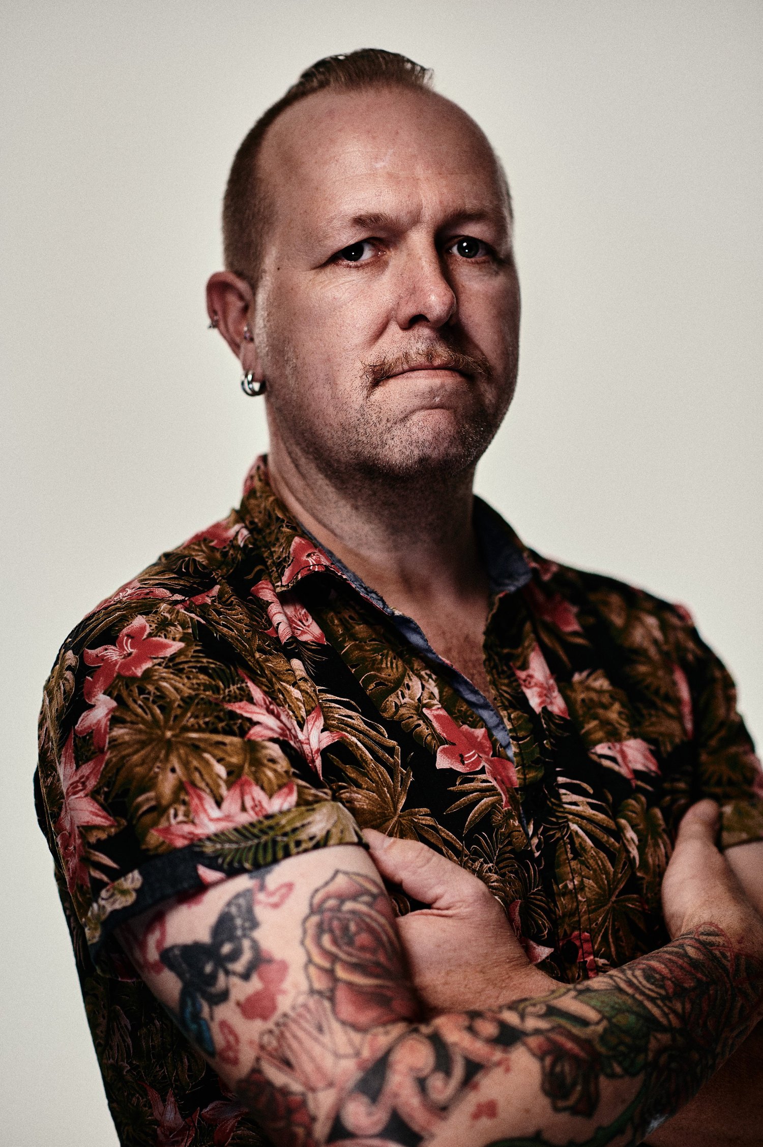 Mann mit tätowiertem Arm, bekleidet mit einem Hawaiihemd, verschränkt die Arme.