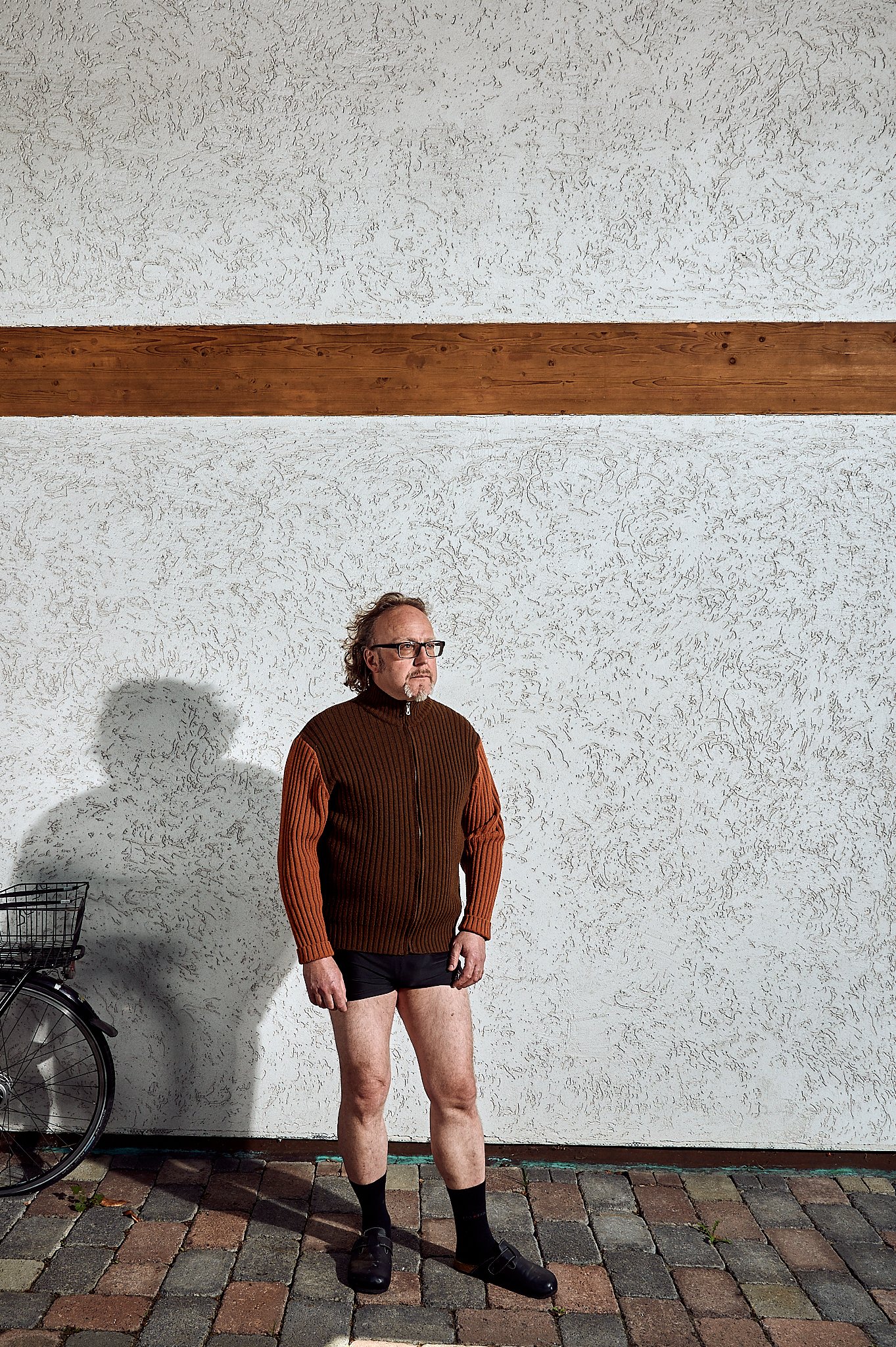Ein Mann steht vor einer strukturierten Wand, trägt einen braunen Pulli und schwarze Shorts. Neben ihm ist ein Fahrrad zu sehen.