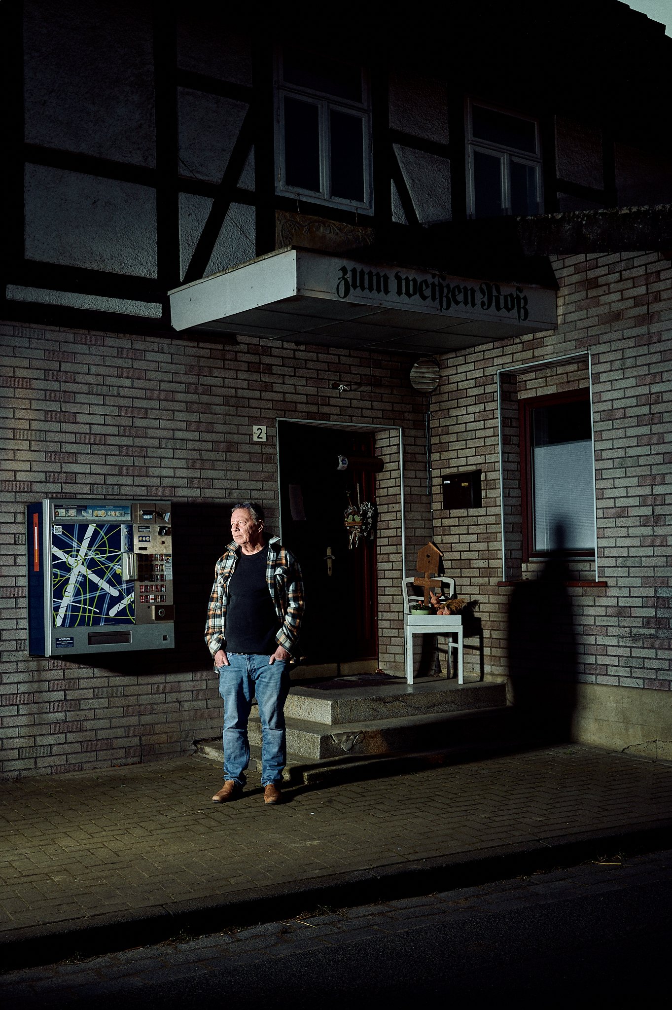Ein Mann steht vor einem Gebäude mit der Aufschrift 'zum weißen Roß'. Das Gebäude hat eine Backsteinfassade und ein Zigarettenautomat ist an der Wand angebracht. Der Mann trägt eine karierte Jacke, Jeans und braune Schuhe. Neben der Eingangstür steht