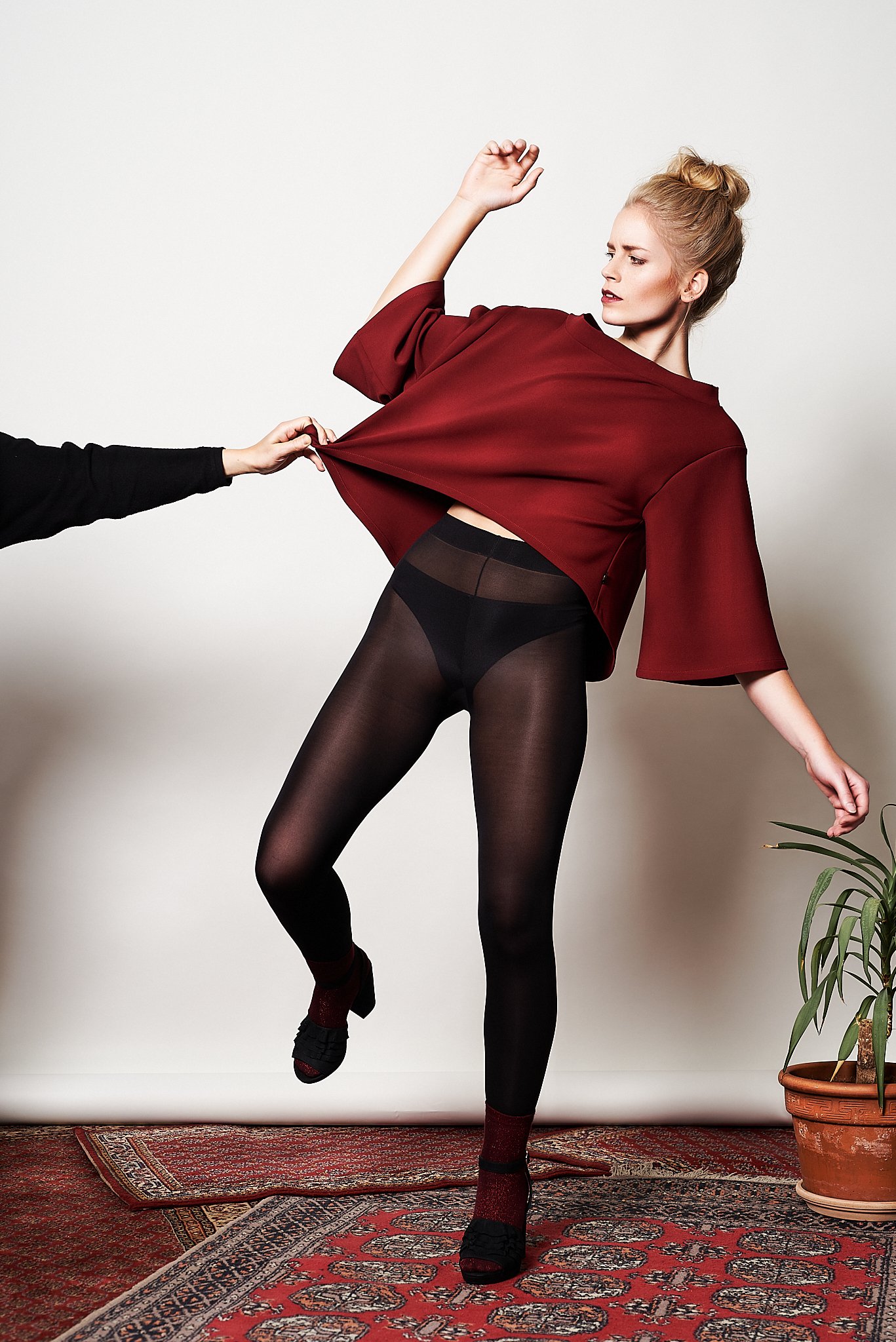 Frau in rotem Oberteil und schwarzer Strumpfhose wird von einer anderen Person am Shirt gezogen, steht auf einem roten Teppich neben einer Pflanze.