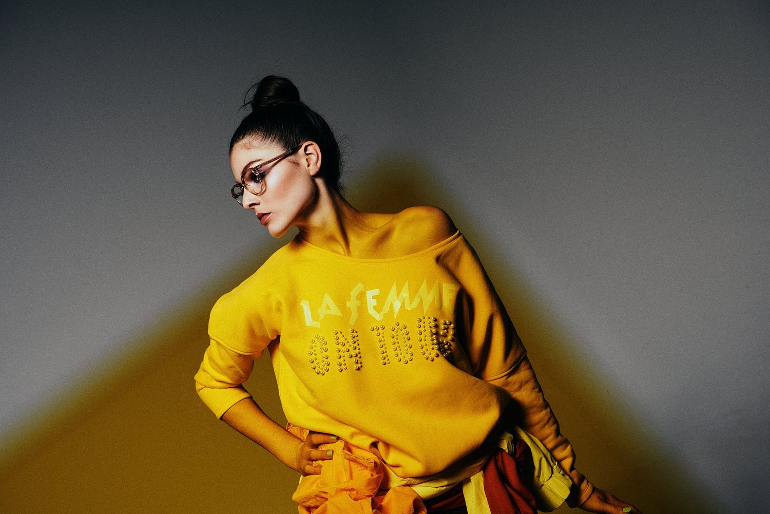 Frau in gelbem Pullover mit "La Femme"-Aufdruck, Brille, hochgesteckter Frisur, steht in Pose vor grauem Hintergrund.