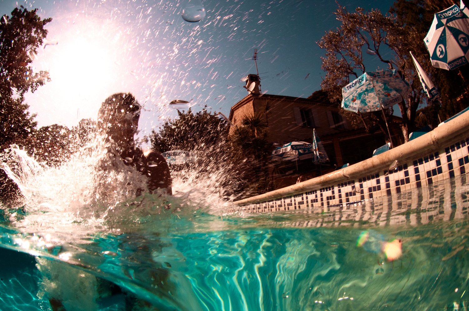 Person im Pool, Wasser spritzt, sonniger Himmel, Bäume und Gebäude im Hintergrund, Sonnenschirme am Poolrand.