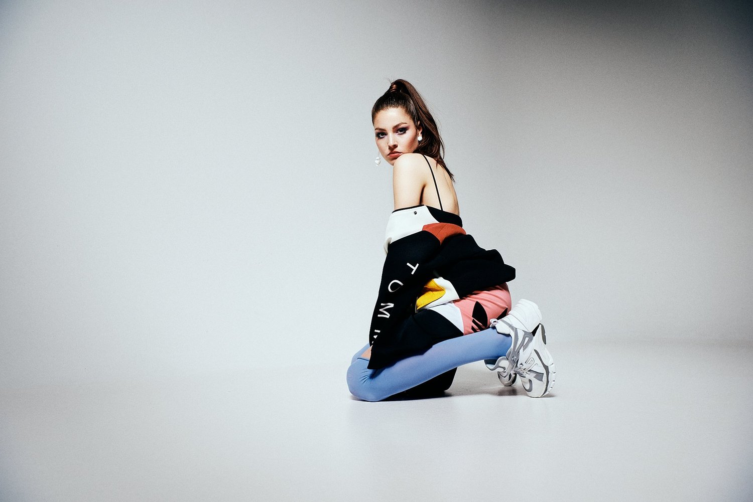 Eine Frau in sportlicher, farbenfroher Kleidung kniet auf dem Boden und schaut zur Seite. Sie trägt weiße Sneakers, eine bunte Jacke und blaue Leggings.