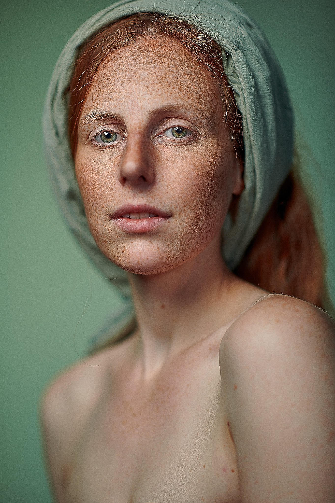 Nahaufnahme einer Person mit roten Haaren und Sommersprossen, die ein grünes Tuch auf dem Kopf trägt, vor einem grünen Hintergrund.