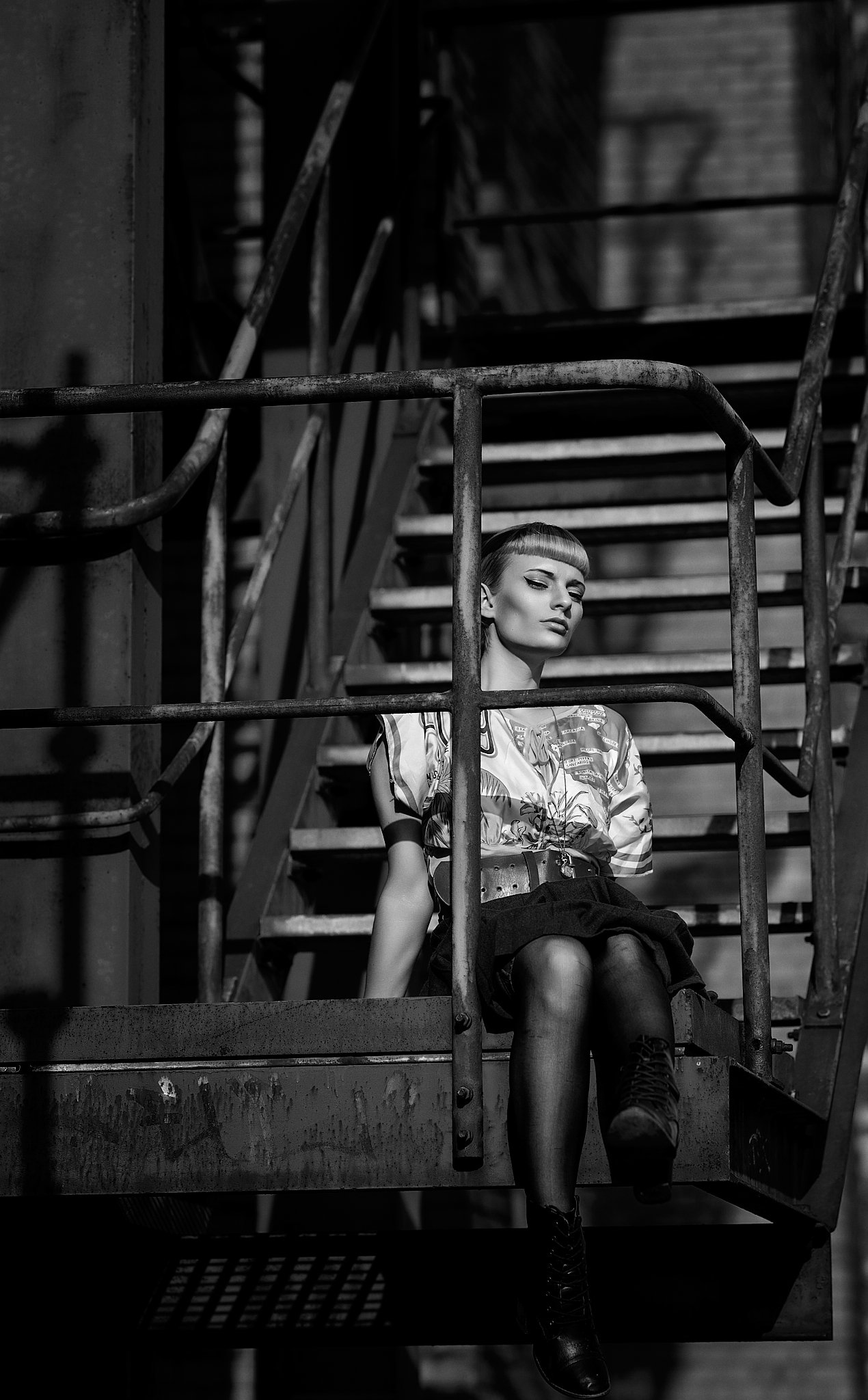 Frau sitzt auf einer metallenen Treppe in Schwarz-Weiß-Fotografie, trägt ein gemustertes Shirt und dunkle Stiefel.