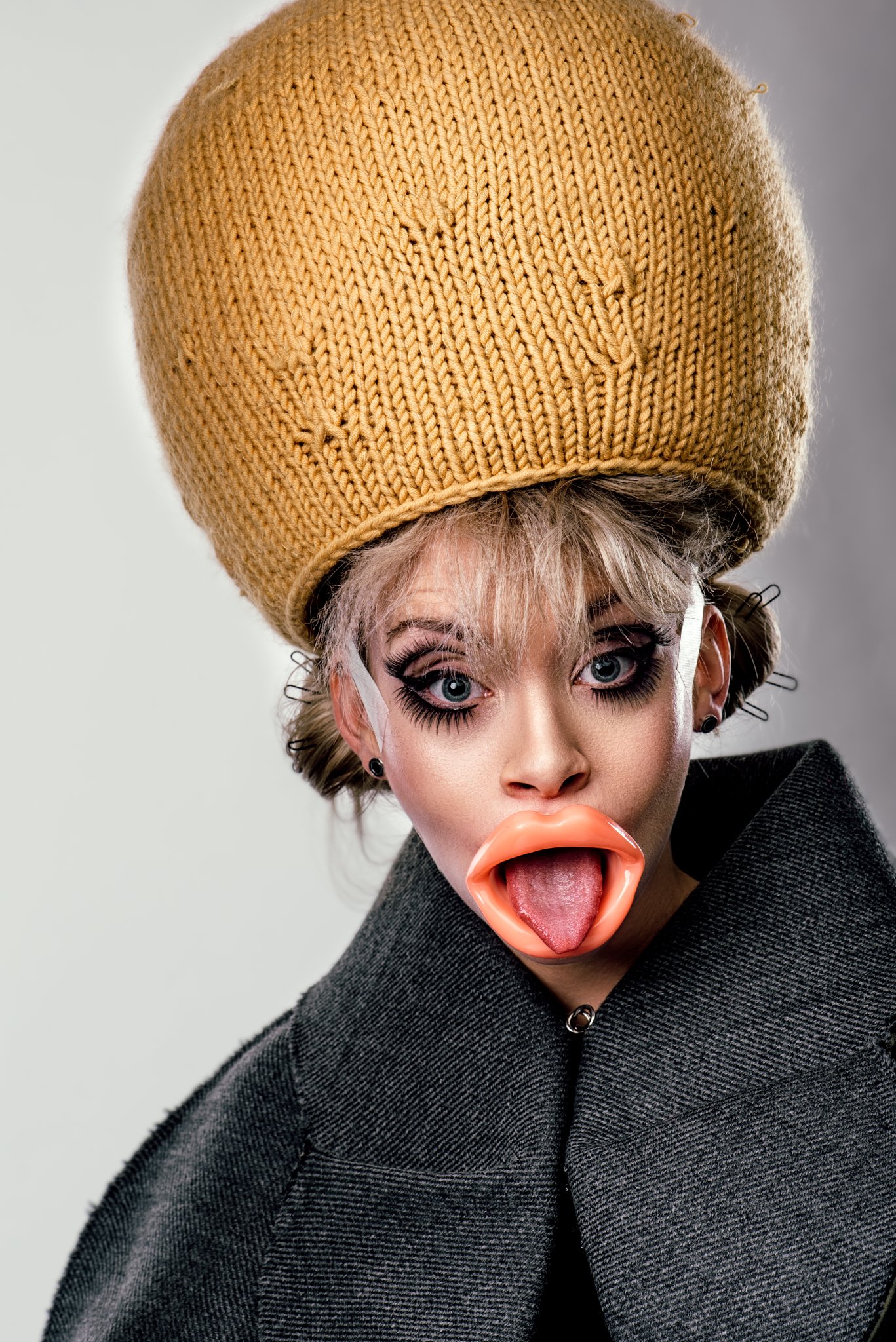 Person mit übergroßem Strickhut, auffälligem Make-up und großen Plastiklippen, die die Zunge herausstreckt, trägt einen dunklen Mantel.