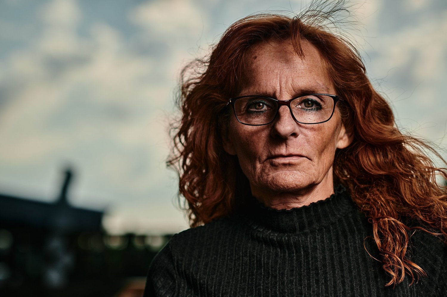 Porträt einer Frau mit Brille und roten Haaren, die ernst in die Kamera schaut. Sie trägt einen dunklen Rollkragenpullover. Der Hintergrund ist unscharf, es scheint draußen zu sein.