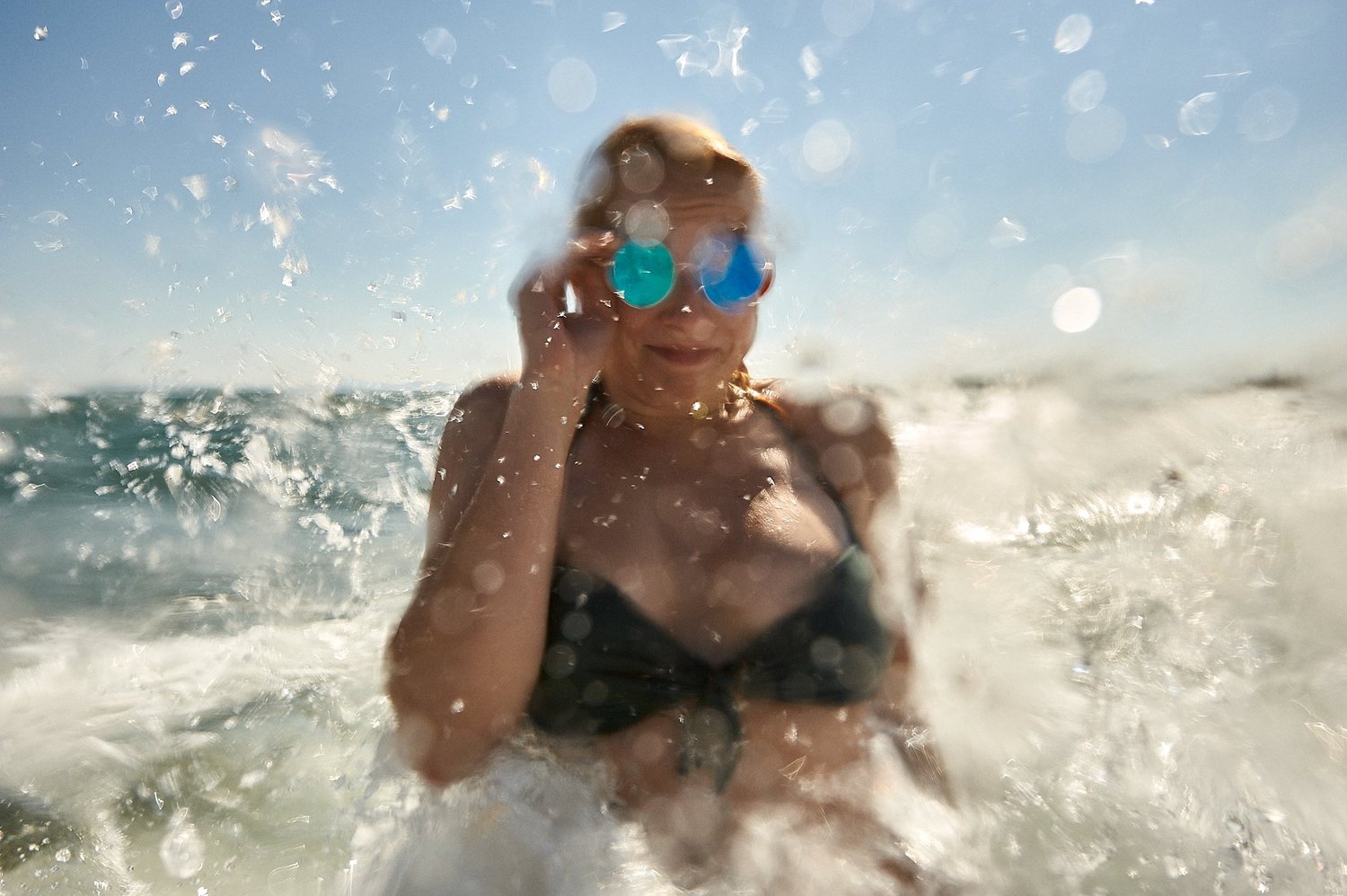 Verschwommene Aufnahme einer Person mit Sonnenbrille am Strand, umgeben von spritzendem Wasser.