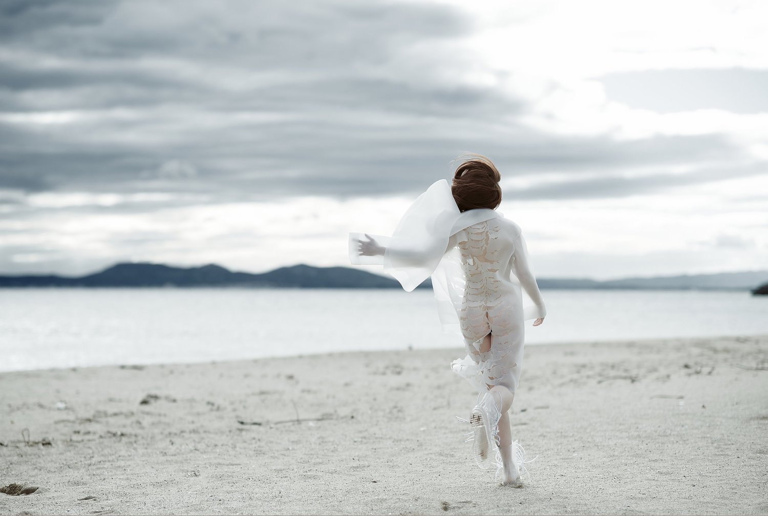 Person am Strand in weißem, durchscheinendem Outfit, läuft Richtung Meer.