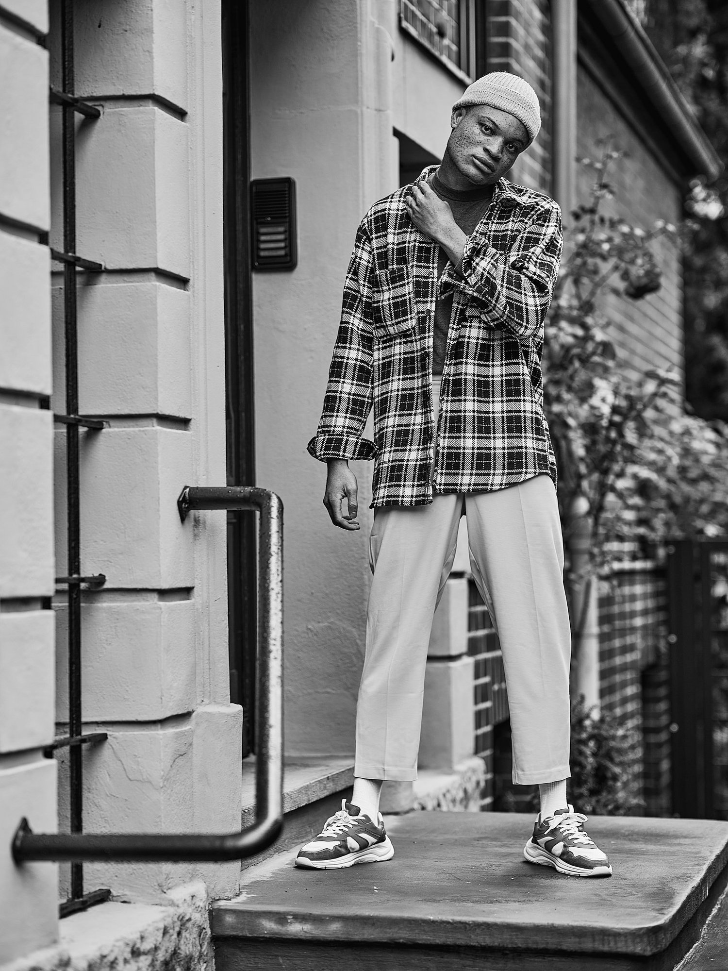 Schwarz-Weiß-Foto einer Person in einem karierten Hemd, Hose und Sneakers auf einer Außentreppe, mit geneigtem Kopf und einer Hand am Hals, vor einem Gebäude stehend.