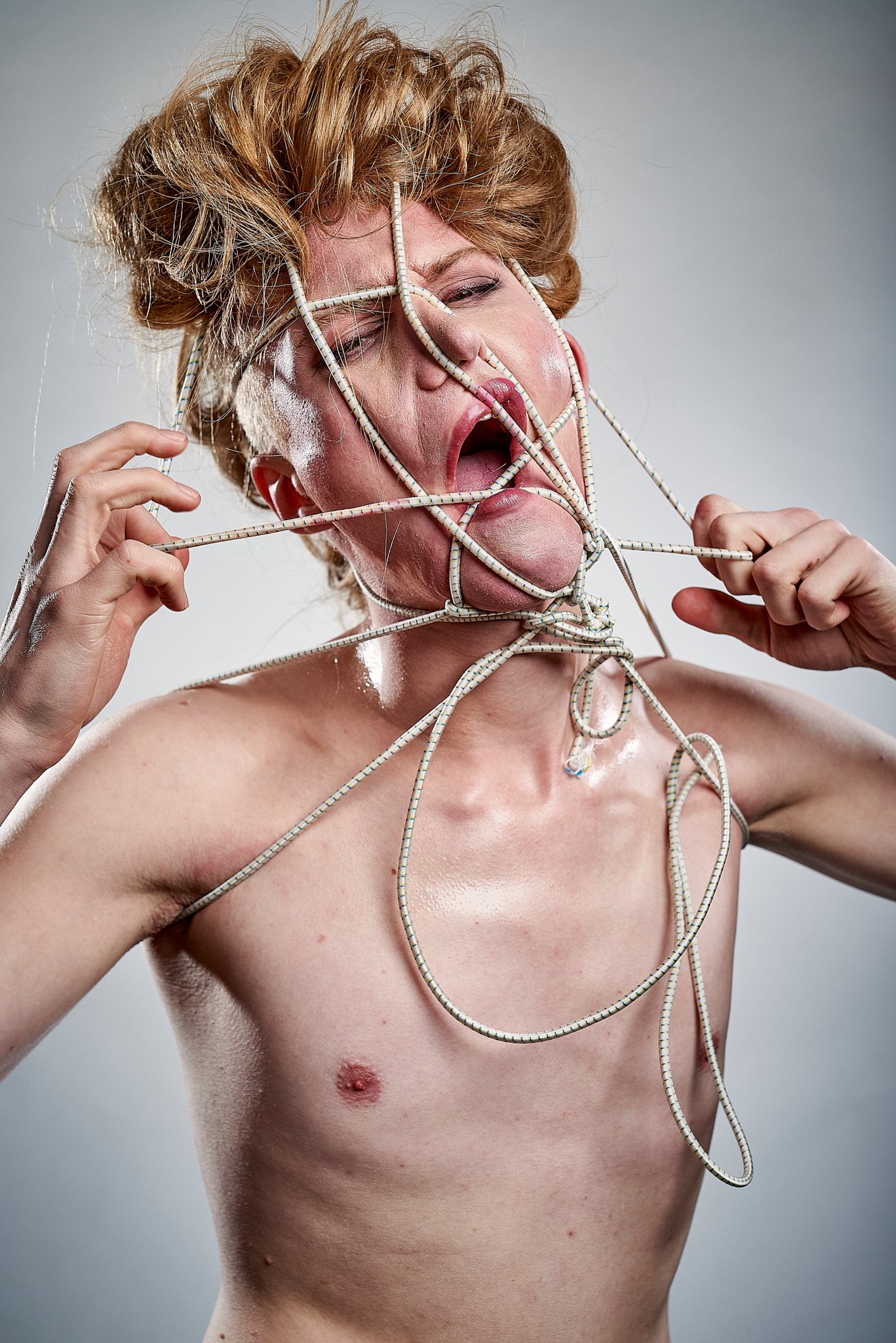 Oberkörper einer Person mit zerrauftem Haar, die mit Seilen um den Kopf gebunden ist, der Mund offen in einem kunstvollen Ausdruck.