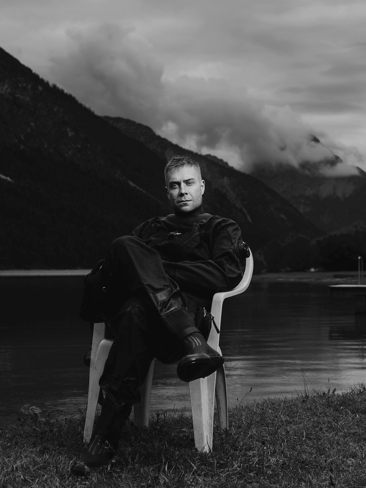 Ein junger Mann in dunkler Kleidung sitzt auf einem weißen Stuhl am Ufer eines Sees mit Bergen und Wolken im Hintergrund, schwarz-weiß Aufnahme. (c Dino Mari, Dino Foto)