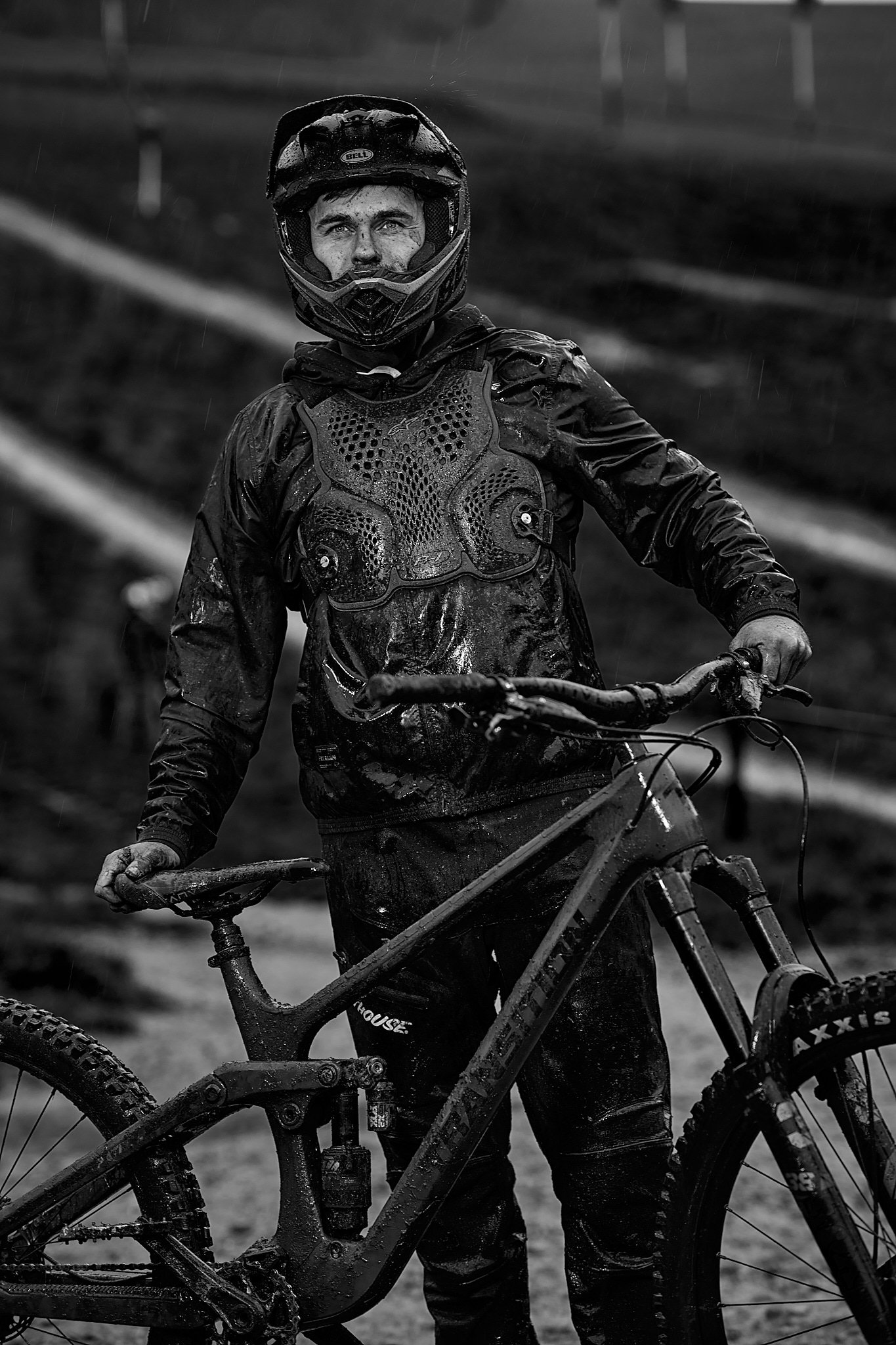 Ein Mountainbiker im Regen, trägt Schutzkleidung und einen Helm, hält ein verschlammtes Fahrrad. Das Schwarz-Weiß-Foto zeigt eine regnerische und schlammige Umgebung, die den Extremsportcharakter unterstreicht.