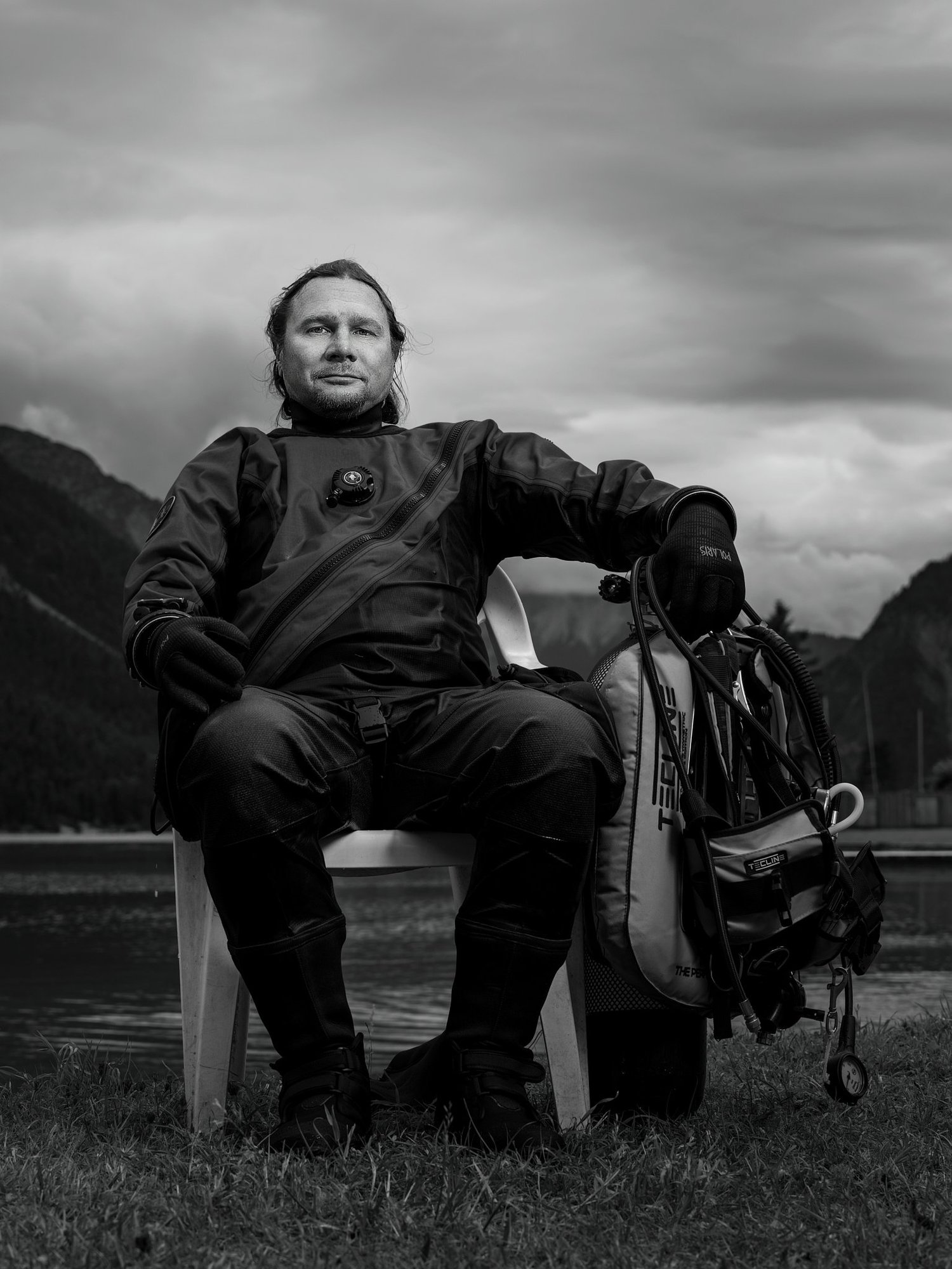 Mann in Outdoor-Ausrüstung, sitzend auf einem Stuhl am See mit Bergen im Hintergrund, schwarz-weiß, Ruhe und Natur. (c Dino Mari, Dino Foto)