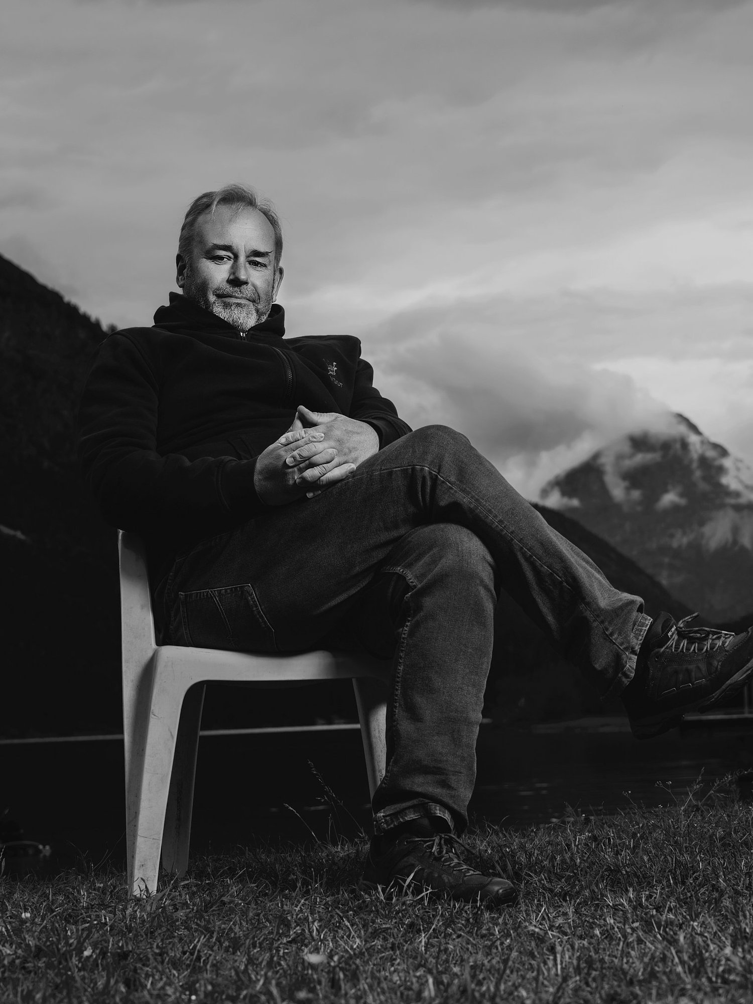 Ein Mann sitzt auf einem weißen Gartenstuhl auf einer Wiese mit Bergen im Hintergrund, im schwarzen und weißen Foto. (c Dino Mari, Dino Foto)