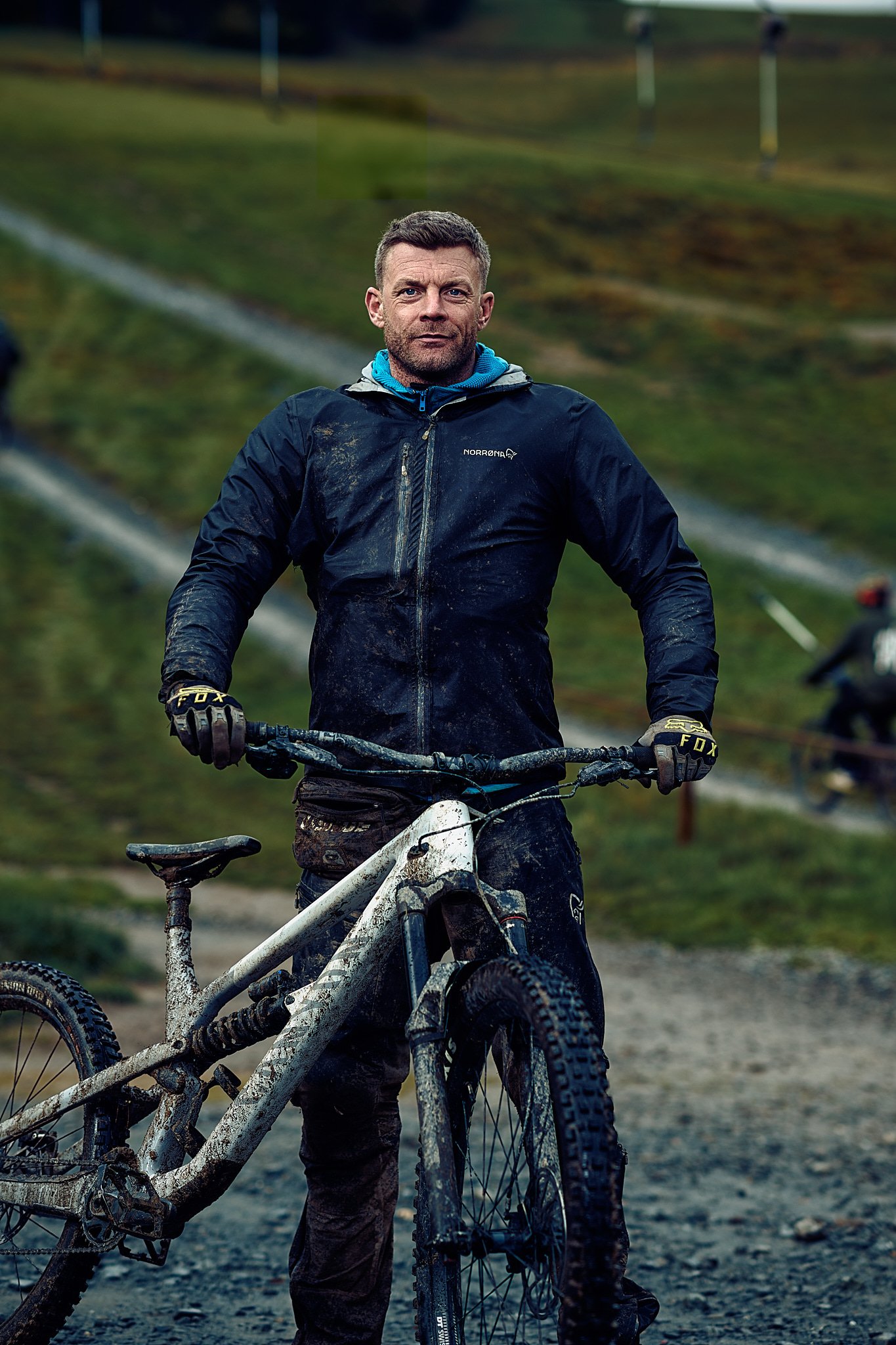 Mann in Outdoor-Kleidung mit einem schlammigen Mountainbike auf einem Feldweg vor einer grünen Landschaft.