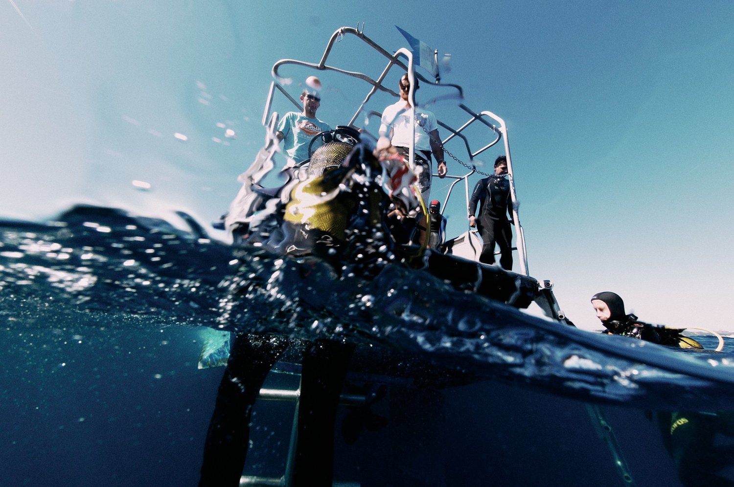 Taucher neben einem Boot im Wasser, teilweise unter Wasser fotografiert. Oben stehen mehrere Personen auf einem kleinen Tauchboot mit Ausrüstung.