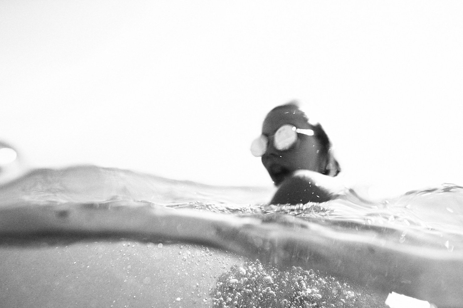 Schwarz-Weiß-Foto einer Person mit Sonnenbrille im Wasser, teils unter Wasser, mit Wellen und sprudelndem Wasser im Vordergrund.