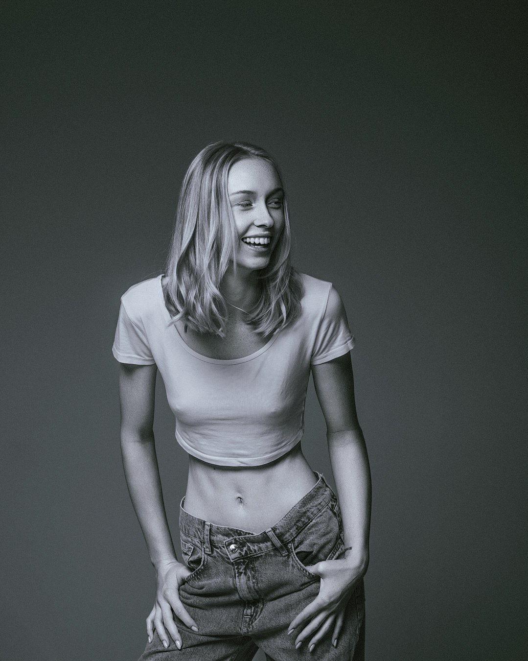 Schwarz-weiß Bild eines lachenden jungen Mädchens mit lockigem, schulterlangem Haar, trägt ein Crop-Top und Jeans, vor dunklem Hintergrund.