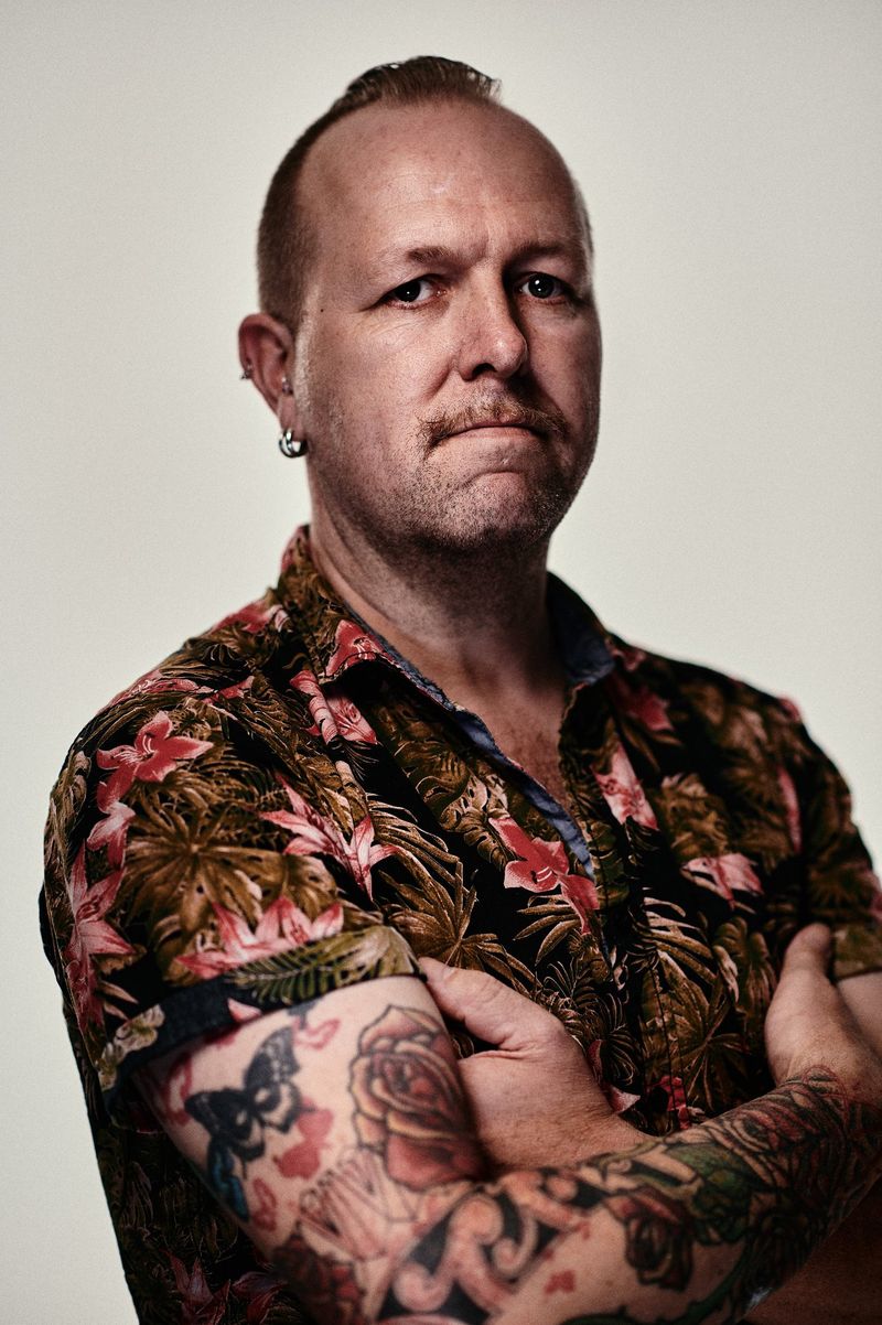Mann mit tätowiertem Arm, bekleidet mit einem Hawaiihemd, verschränkt die Arme.