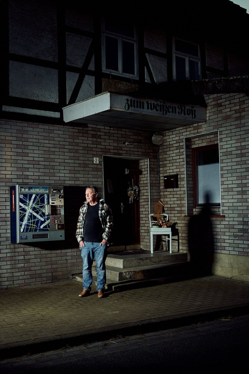 Ein Mann steht vor einem Gebäude mit der Aufschrift 'zum weißen Roß'. Das Gebäude hat eine Backsteinfassade und ein Zigarettenautomat ist an der Wand angebracht. Der Mann trägt eine karierte Jacke, Jeans und braune Schuhe. Neben der Eingangstür steht