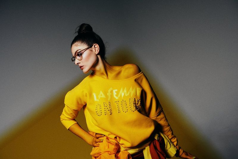 Frau in gelbem Pullover mit "La Femme"-Aufdruck, Brille, hochgesteckter Frisur, steht in Pose vor grauem Hintergrund.