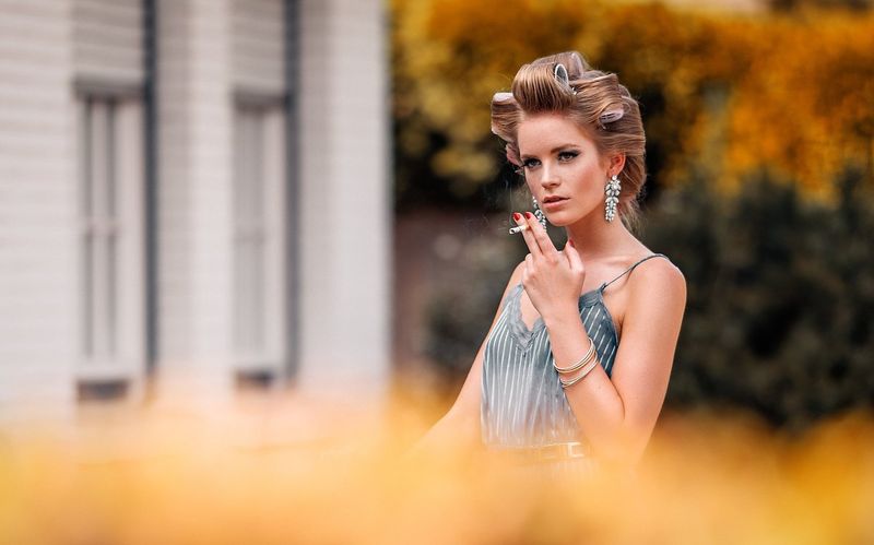 Eine Frau mit retro Frisur und großen Ohrringen hält eine Zigarette. Sie trägt ein elegantes Kleid und steht vor einer verschwommenen Kulisse."}