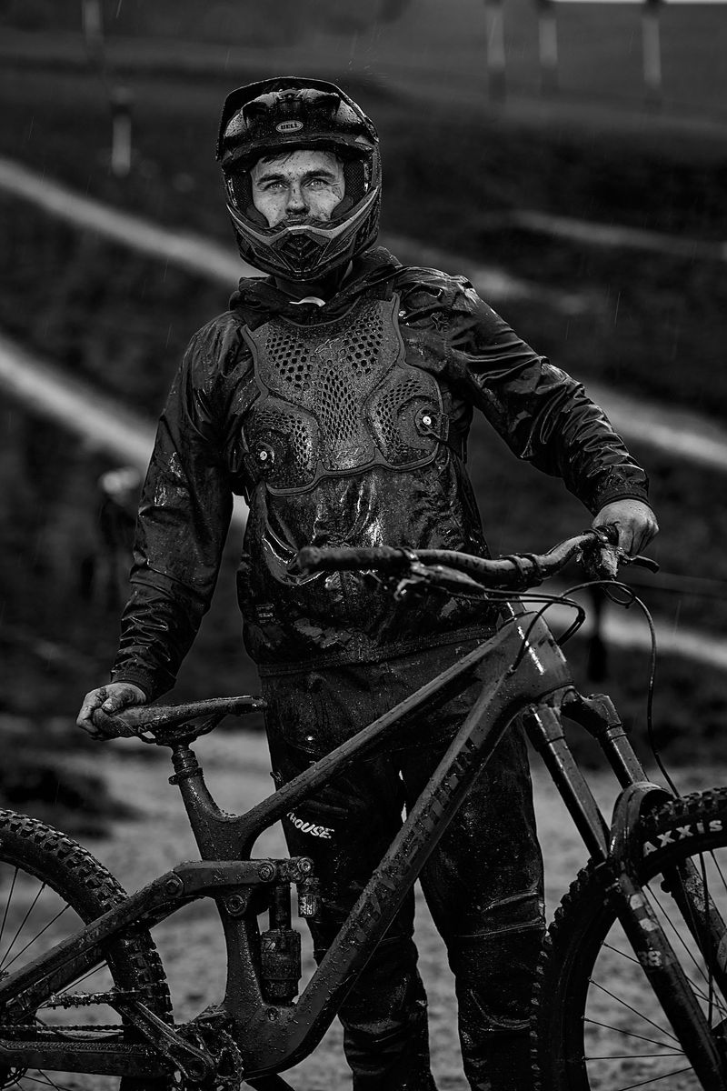 Ein Mountainbiker im Regen, trägt Schutzkleidung und einen Helm, hält ein verschlammtes Fahrrad. Das Schwarz-Weiß-Foto zeigt eine regnerische und schlammige Umgebung, die den Extremsportcharakter unterstreicht.