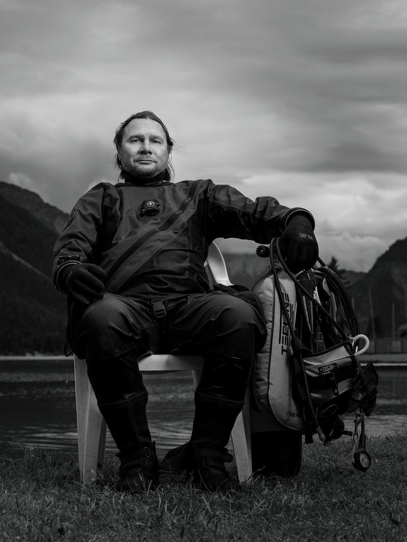 Mann in Outdoor-Ausrüstung, sitzend auf einem Stuhl am See mit Bergen im Hintergrund, schwarz-weiß, Ruhe und Natur. (c Dino Mari, Dino Foto)