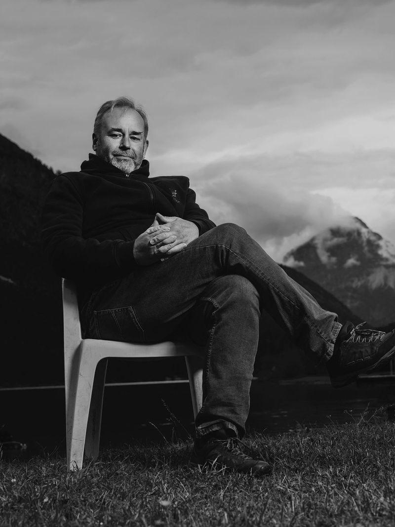 Ein Mann sitzt auf einem weißen Gartenstuhl auf einer Wiese mit Bergen im Hintergrund, im schwarzen und weißen Foto. (c Dino Mari, Dino Foto)