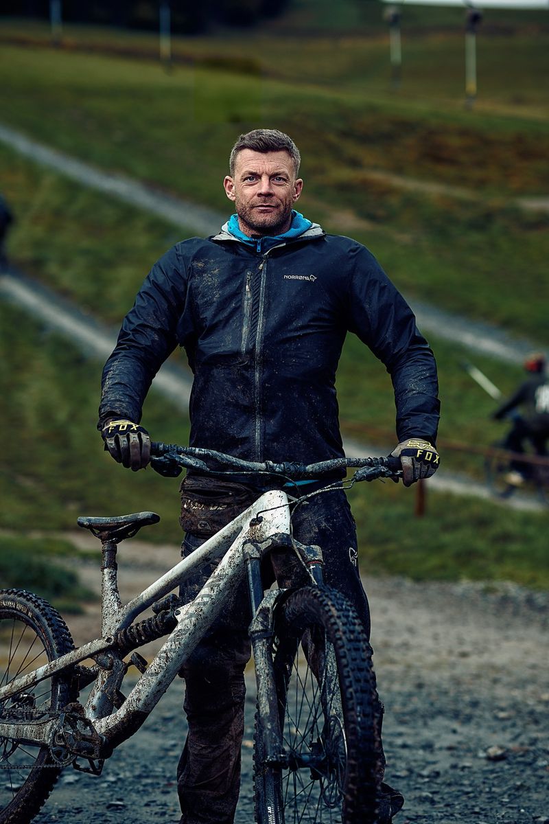 Mann in Outdoor-Kleidung mit einem schlammigen Mountainbike auf einem Feldweg vor einer grünen Landschaft.