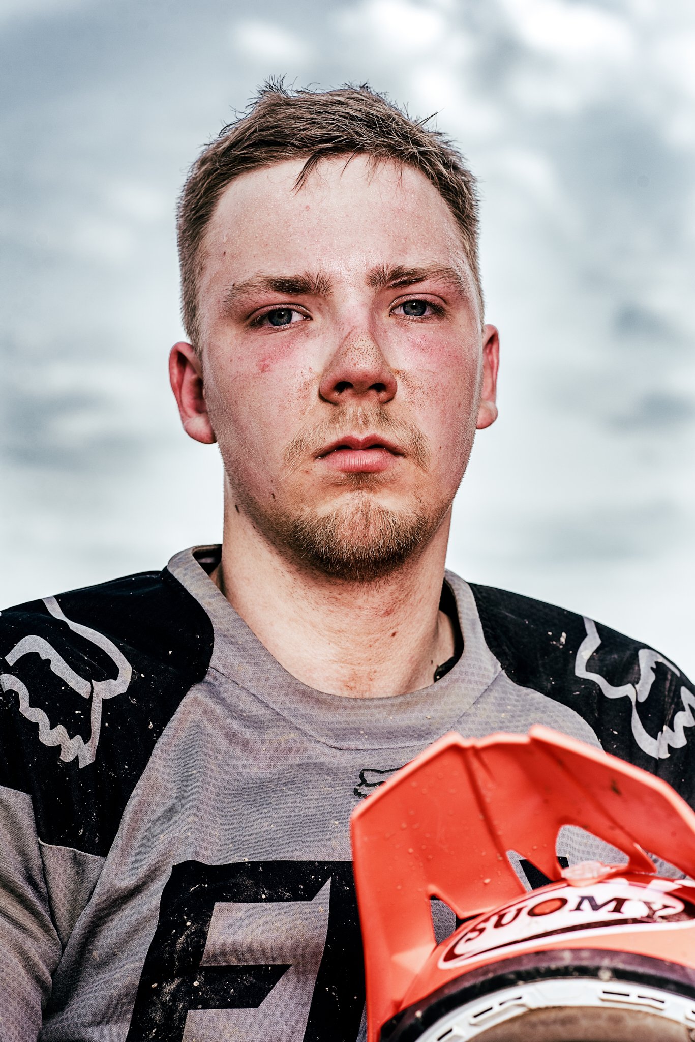 Motocross-Fahrer in Schutzkleidung mit Helm, ernster Gesichtsausdruck, bewölkter Himmel im Hintergrund.