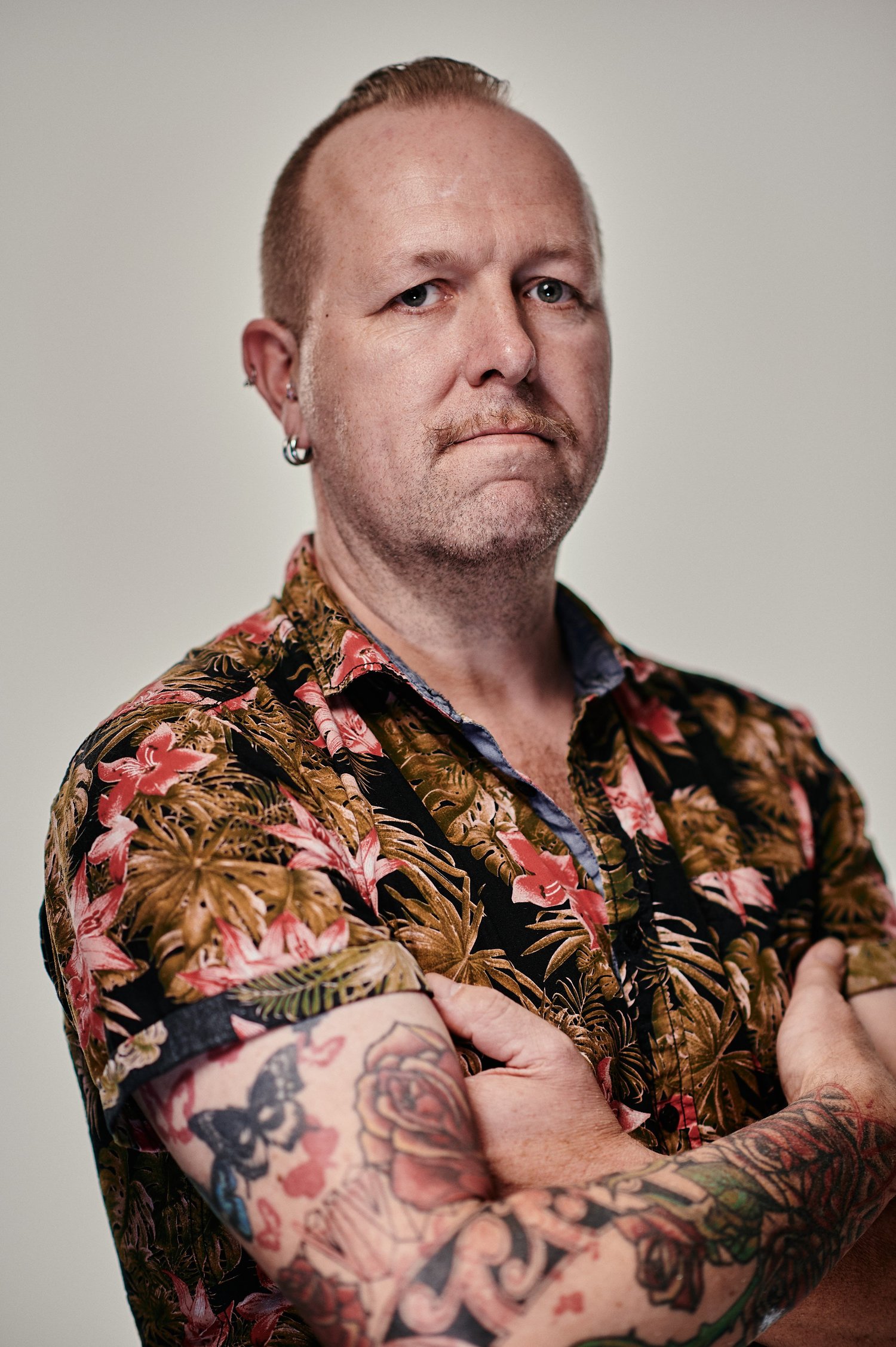 Nahaufnahme eines Mannes mit verschränkten Armen, einem bunten Hemd mit Blumenmuster und Tätowierungen auf dem Arm.