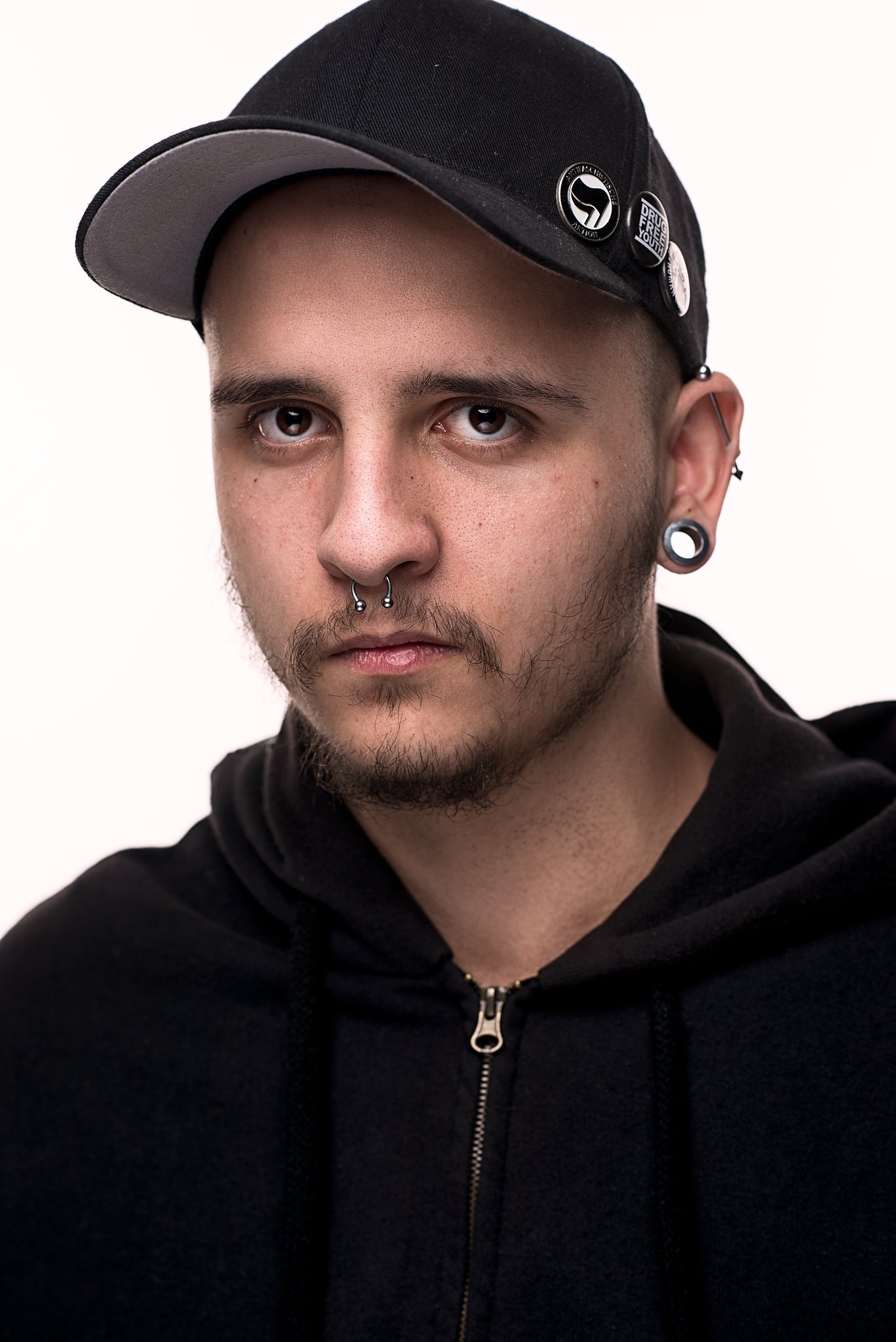 Nahaufnahme eines Mannes mit Piercings und einer schwarzen Kappe, verziert mit Ansteckern, in einem schwarzen Kapuzenpullover.