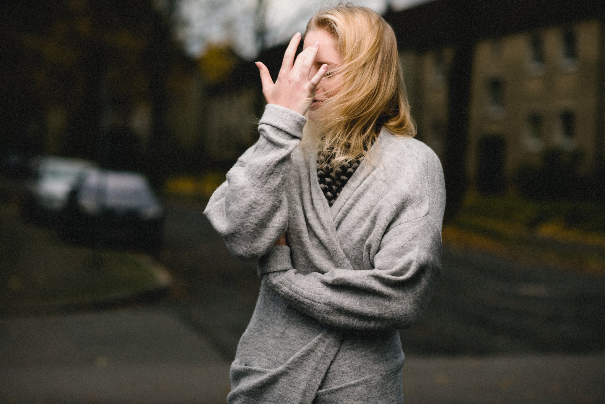 Eine Frau mit blonden Haaren, die ein graues Outfit trägt, steht auf einer Straße und bedeckt ihr Gesicht teilweise mit der Hand.
