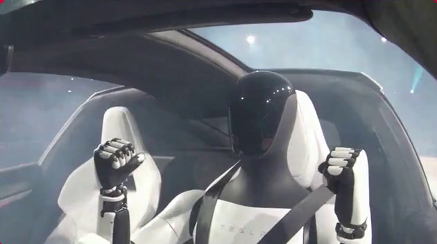 Ein humanoider Roboter mit schwarzem, glänzendem Kopf und der Aufschrift ‚TESLA‘ auf dem Rumpf sitzt angeschnallt auf einem weißen Sitz in einem Fahrzeug und hebt beide Fäuste.