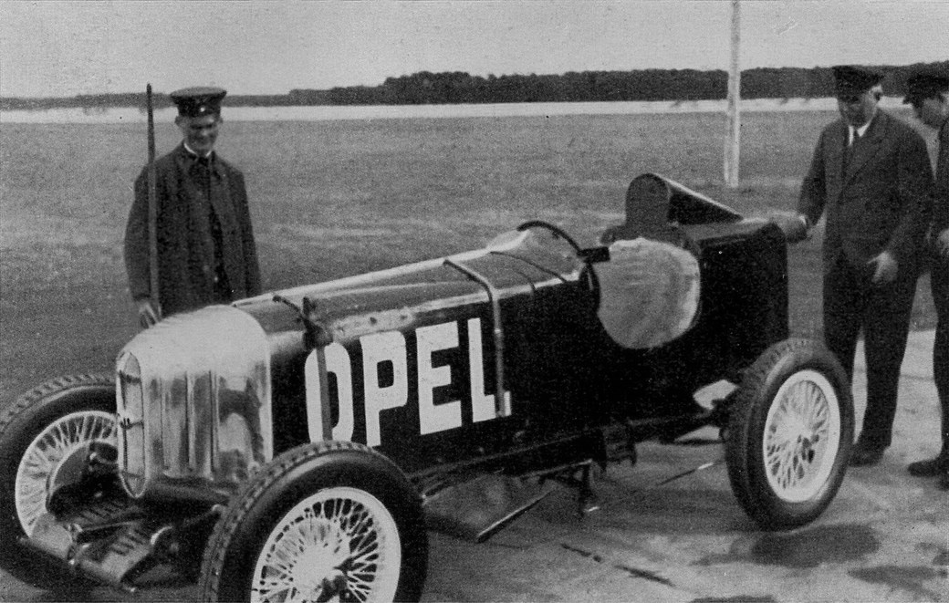 Schwarzweißfoto von 1928: der schwarze Opel-Raketenwagen RAK 1 mit weißem OPEL-Schriftzug auf einem Flugfeld, daneben drei Männer in Uniform und Anzug.